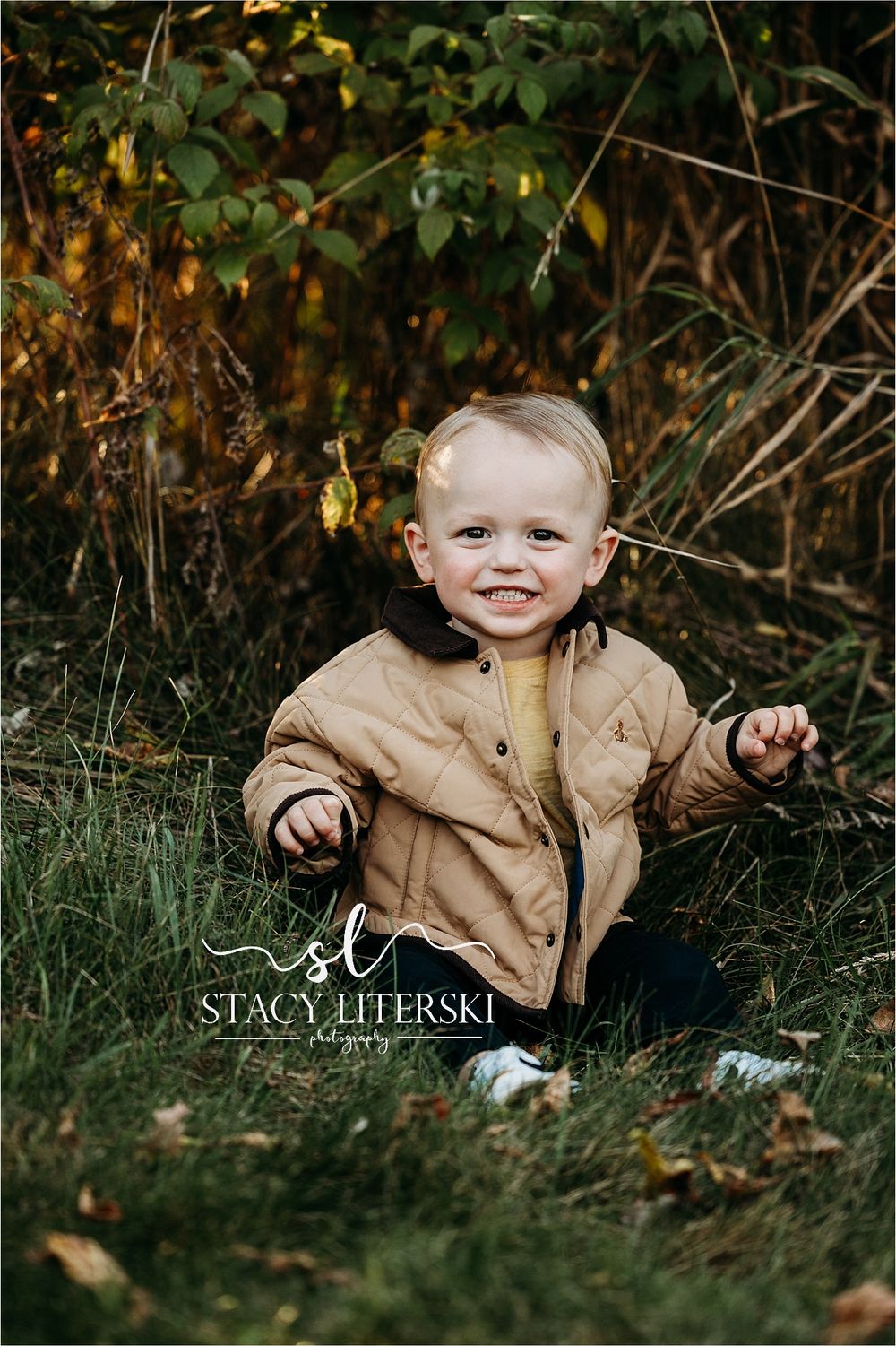 Smiling toddler in a tan quilted jacket sitting in green grass with autumn foliage in the background.