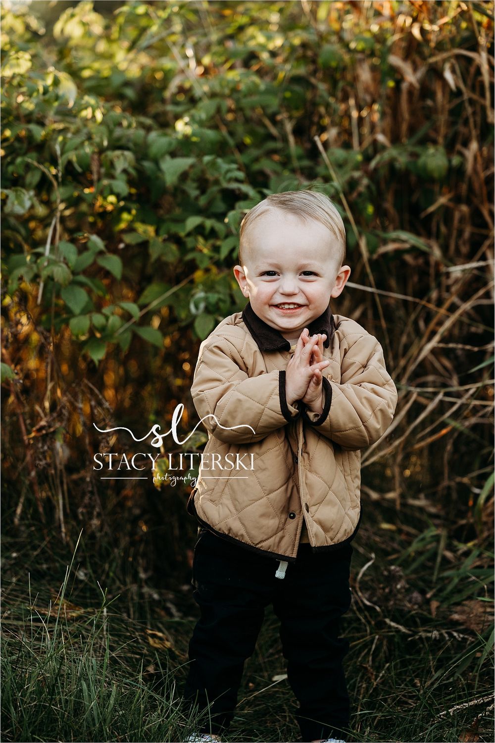 Smiling toddler boy in tan quilted jacket clapping hands outdoors among green foliage in fall portrait.