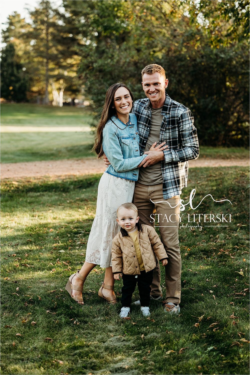 Family of three posing outdoors in a park, parents smiling with toddler in tan jacket, fall family portrait photography.