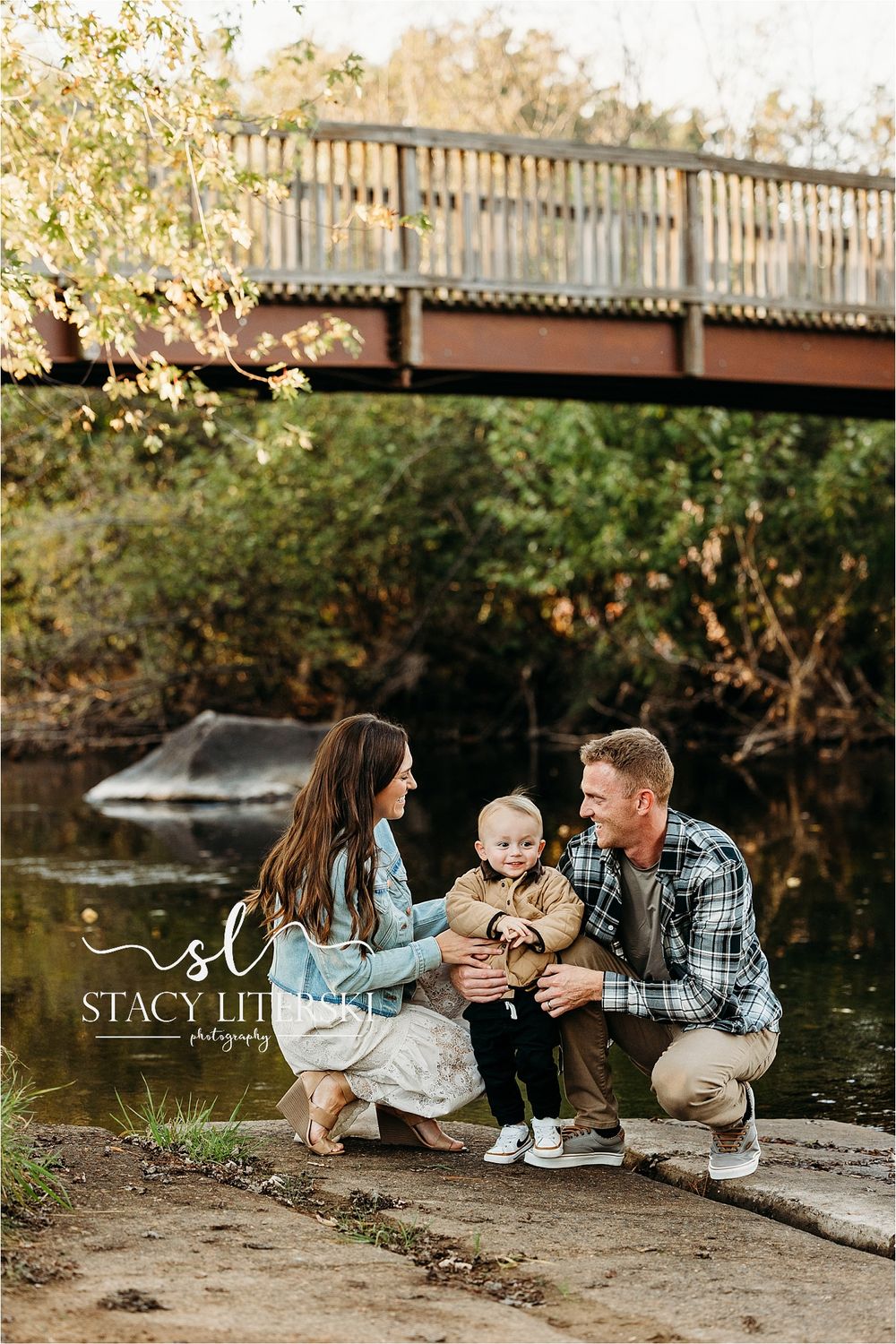 Family of three poses by a creek with a wooden bridge backdrop during an autumn outdoor photo session.