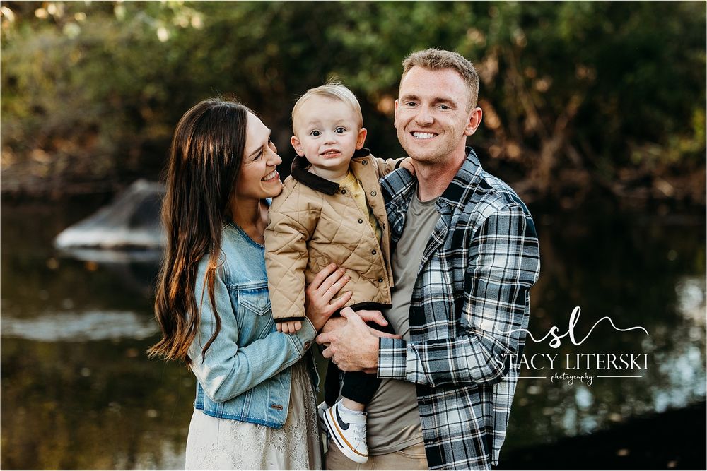 A smiling young family with a toddler boy poses outdoors near a creek in a warm, natural setting.