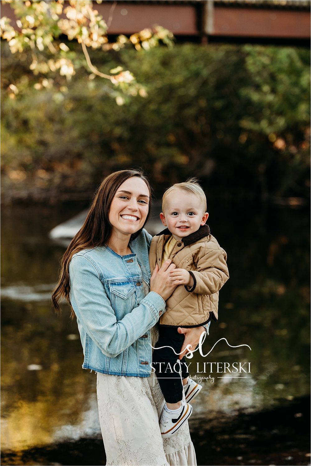 Smiling mother holds her toddler boy outdoors near a creek in a warm autumn family portrait session.