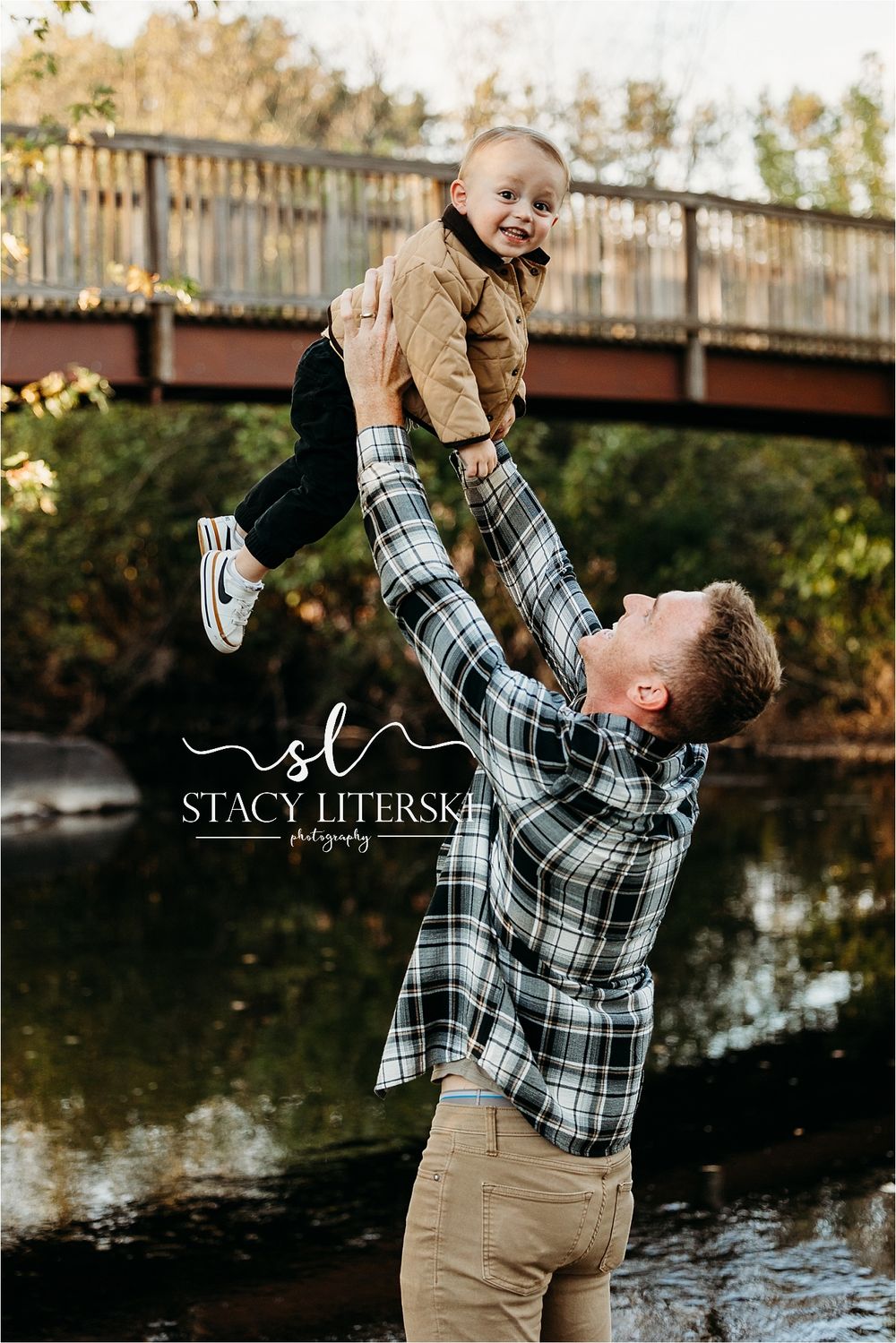 Father joyfully lifting smiling toddler overhead outdoors near a bridge, captured by Stacy Literski Photography.