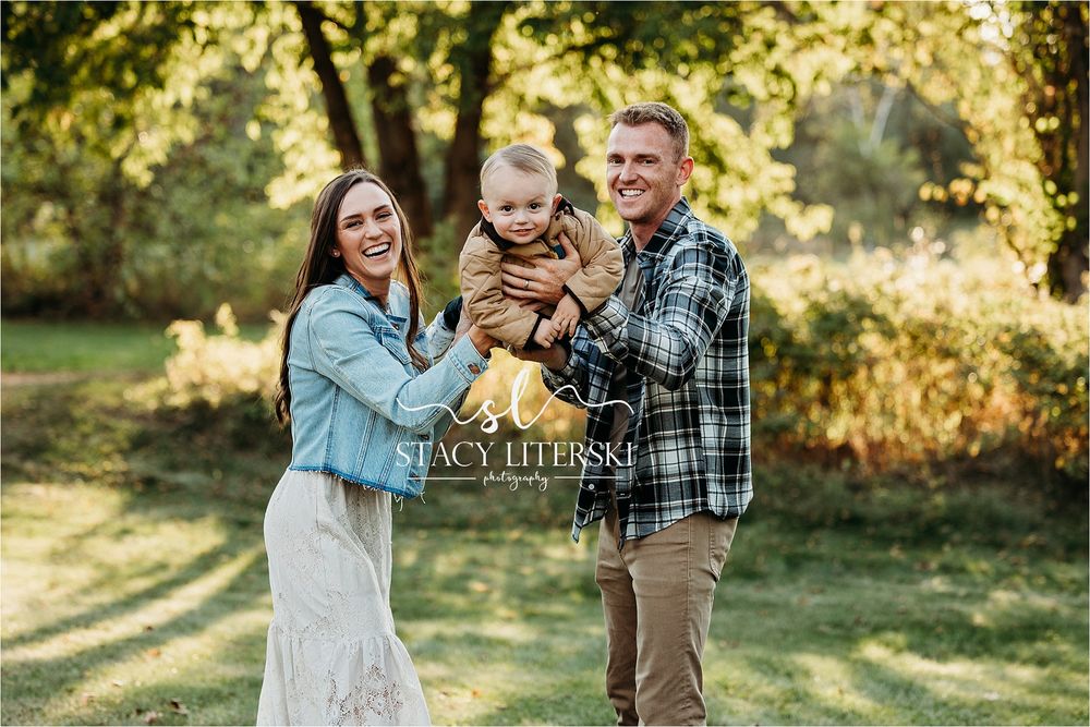 Happy family of three posing outdoors in a lush green park, parents smiling and holding their baby.