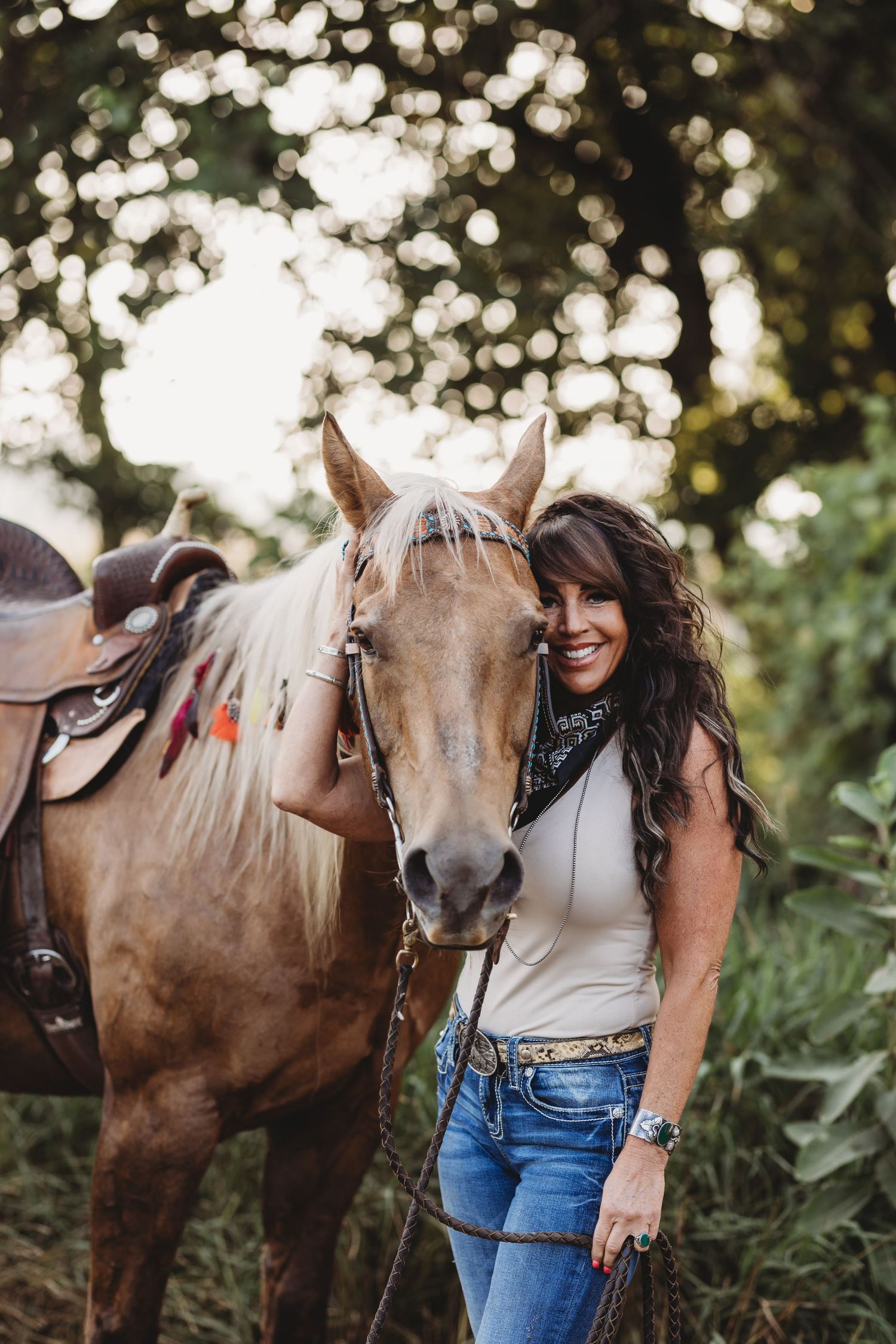 Smiling woman in jeans and bandana stands beside a saddled palomino horse outdoors among green trees.