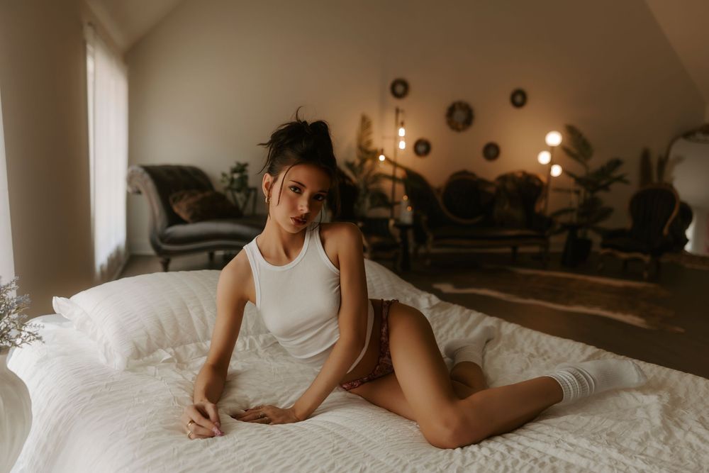 A young woman in a white tank top and brown shorts sits gracefully on a white bed in a cozy, warmly lit bedroom.
