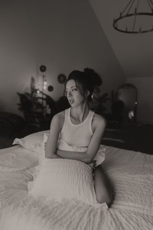 Black and white photo of a young woman sitting on a bed in a white tank top and skirt, looking to the side.