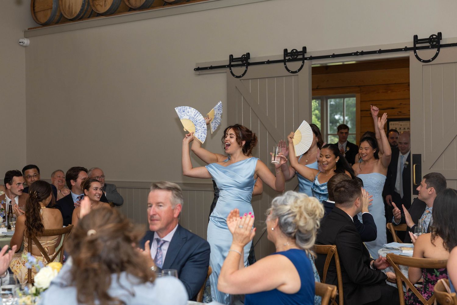 Bridesmaids in blue dresses wave fans while guests celebrate at a rustic wedding reception indoors.