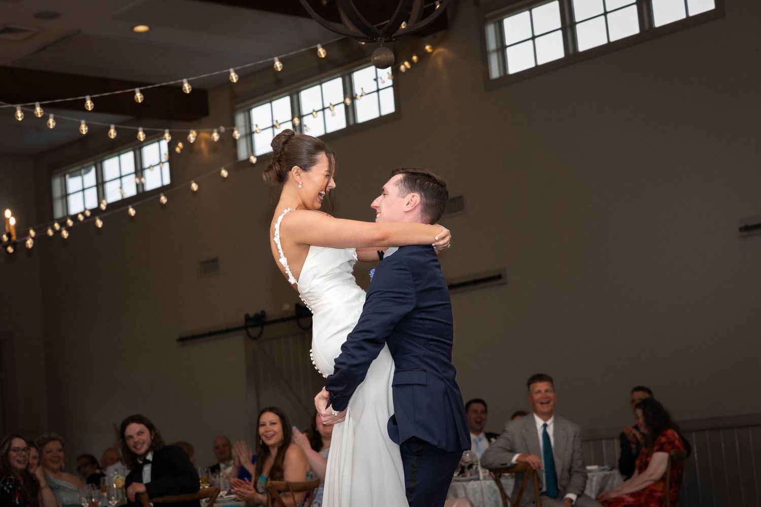 Groom lifts bride during first dance at elegant wedding reception with string lights.