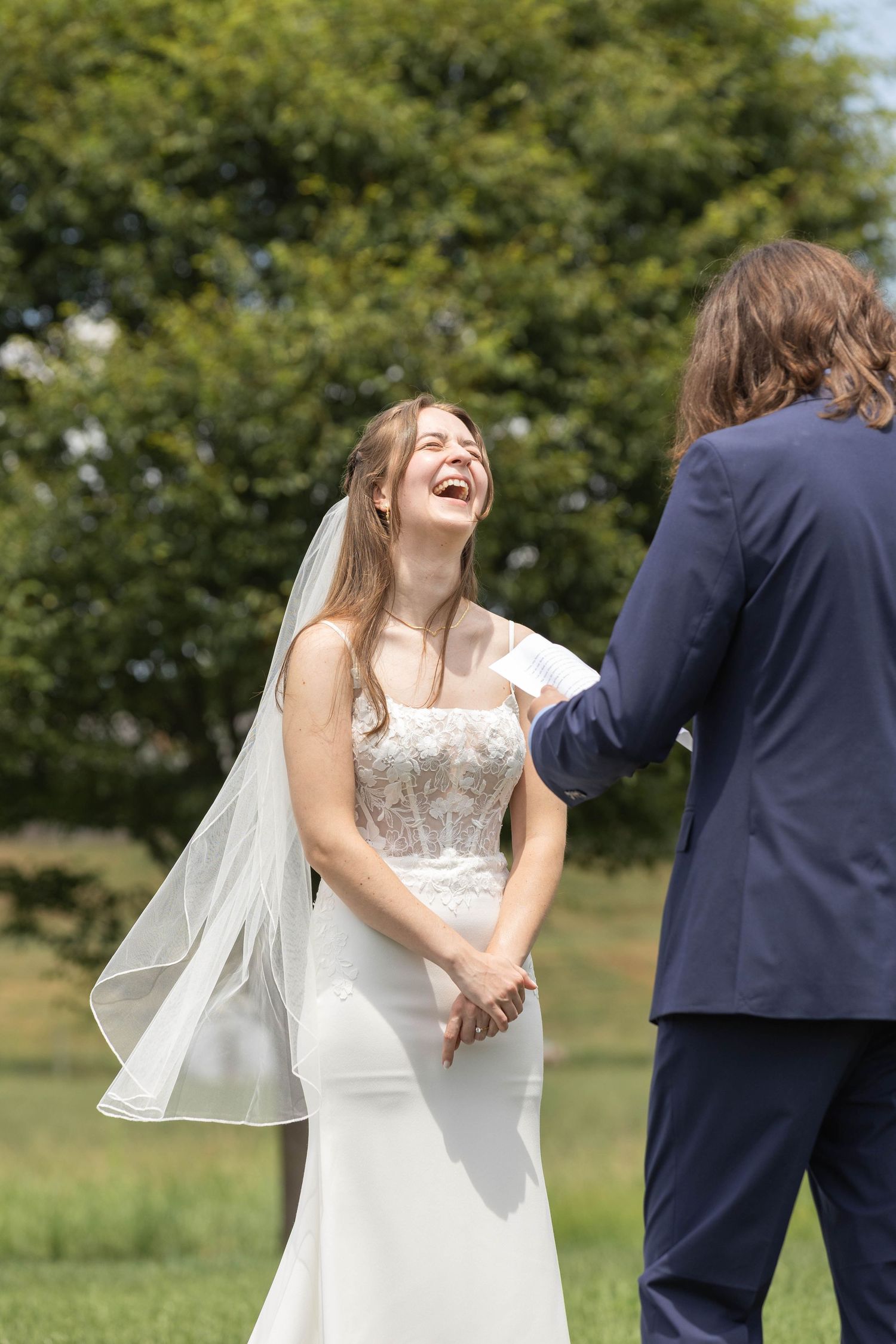 Laughing bride in white lace gown and veil holds hands with groom in navy suit outdoors on sunny wedding day.