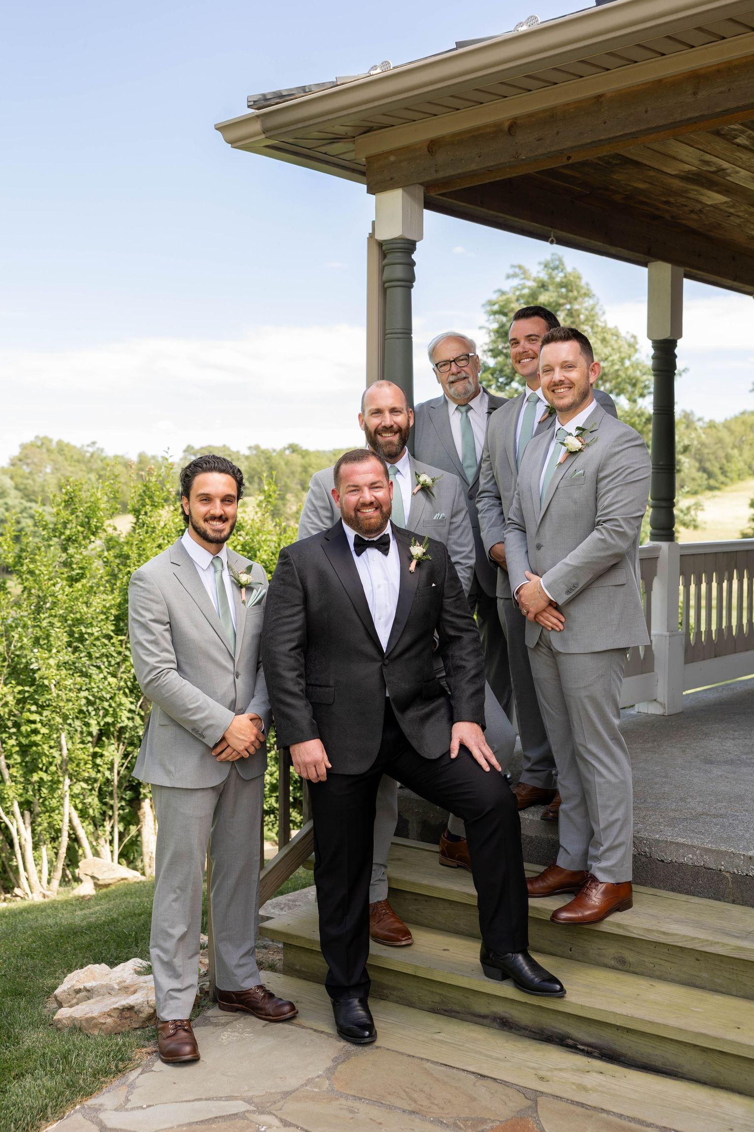 Groom in black tuxedo poses with groomsmen in gray suits on wooden steps at an outdoor wedding venue.