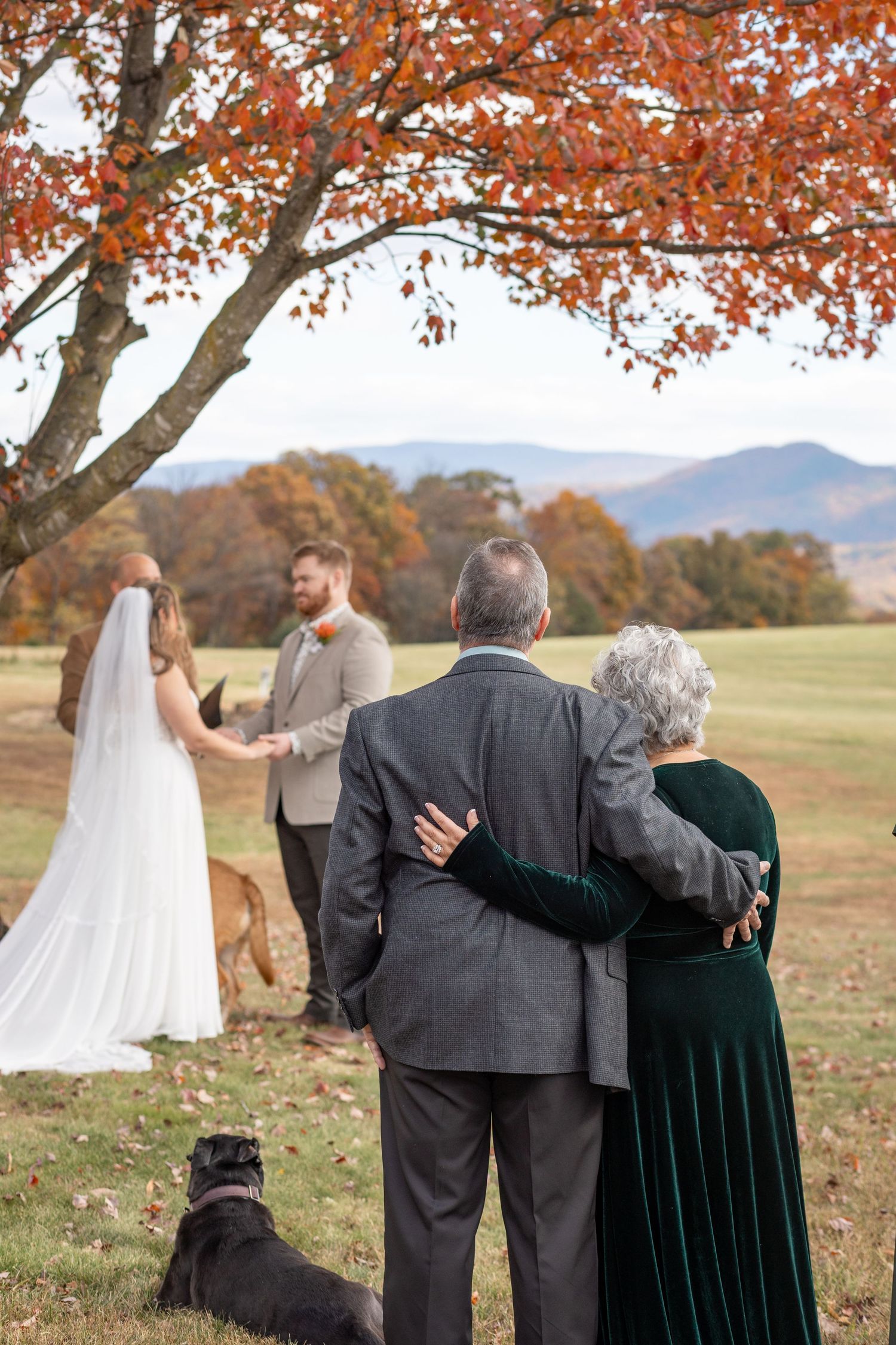Elderly couple embracing while watching outdoor autumn wedding ceremony with mountains in background.