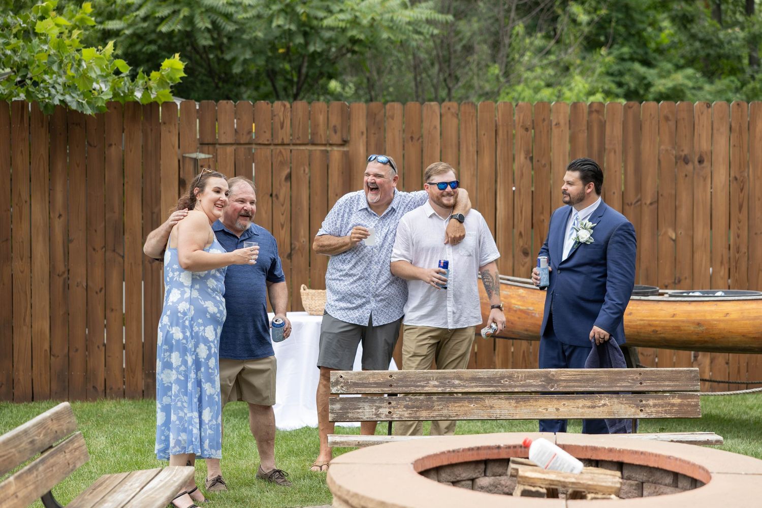 Group of friends laughing and celebrating outdoors near a fire pit with a wooden fence backdrop.