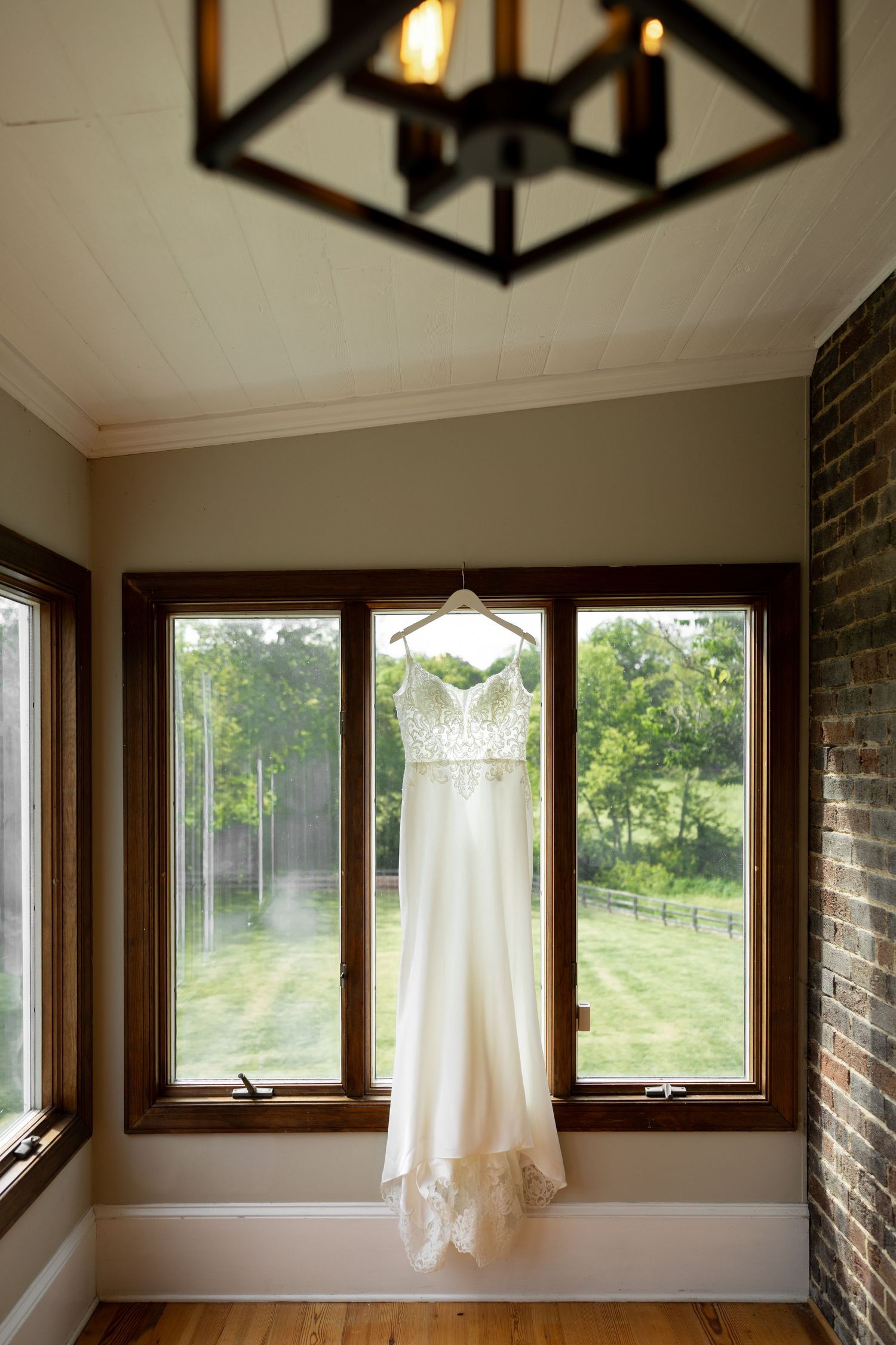 White wedding dress hanging on a hanger in front of large windows with a rustic chandelier above in a charming venue.