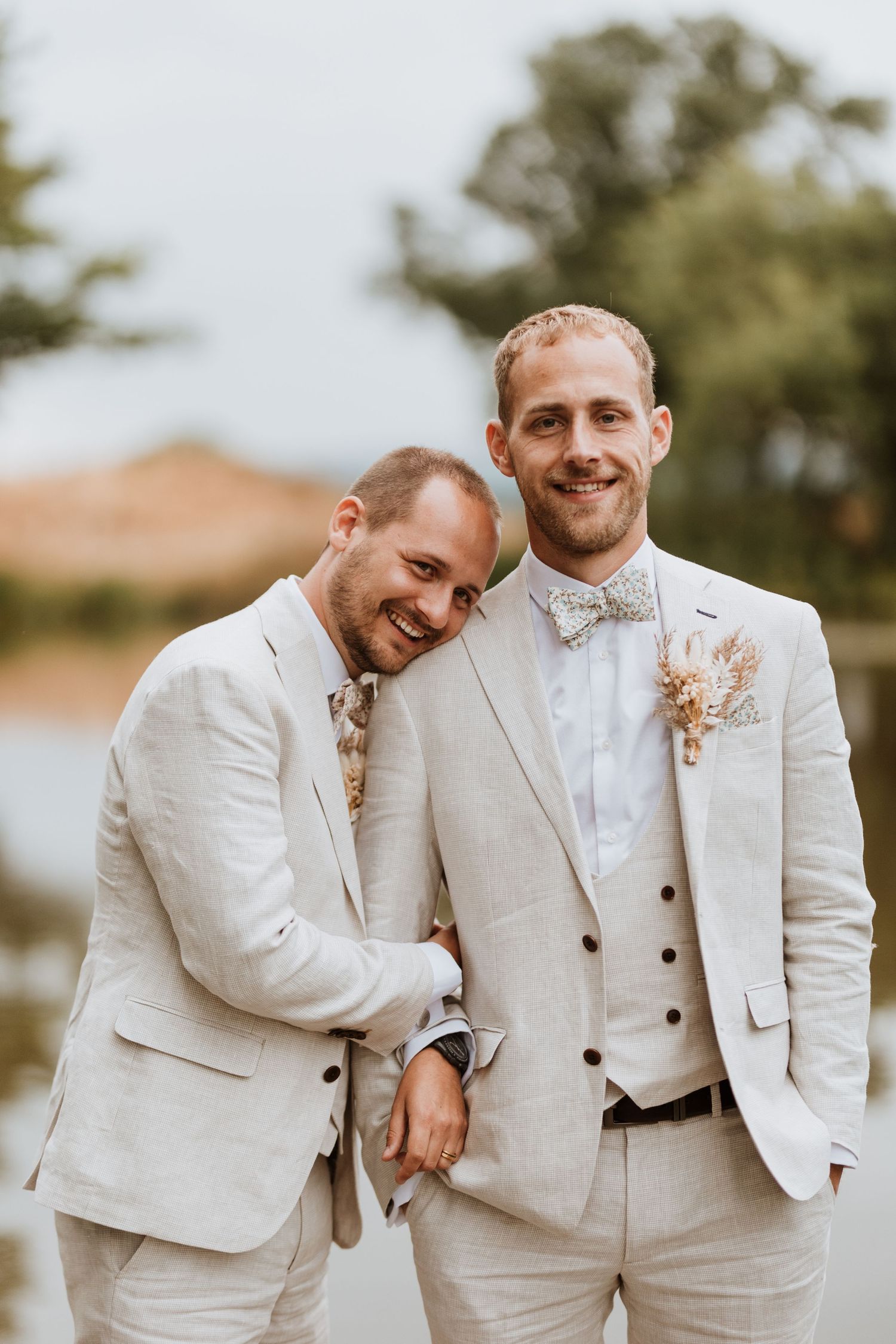 Two grooms in matching cream linen suits with floral boutonnieres smile and embrace outdoors near a lake at Moat Hall Barns