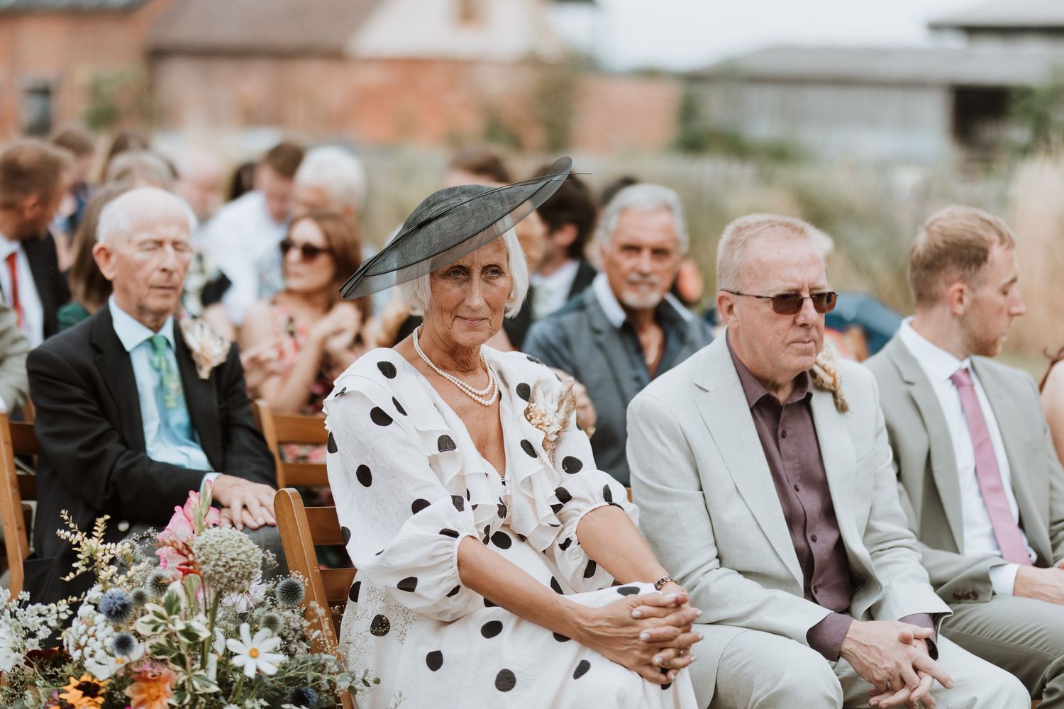 Wedding guests seated outdoors, woman in polka dot dress and black hat, man in sunglasses at a summer ceremony.