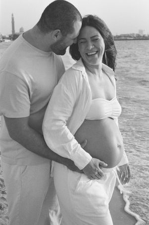 Happy pregnant couple embracing on beach, woman laughing in white outfit, black and white maternity photo.