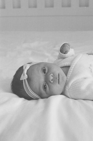 Black and white photo of a newborn baby girl wearing a bow headband, lying on white bedding.