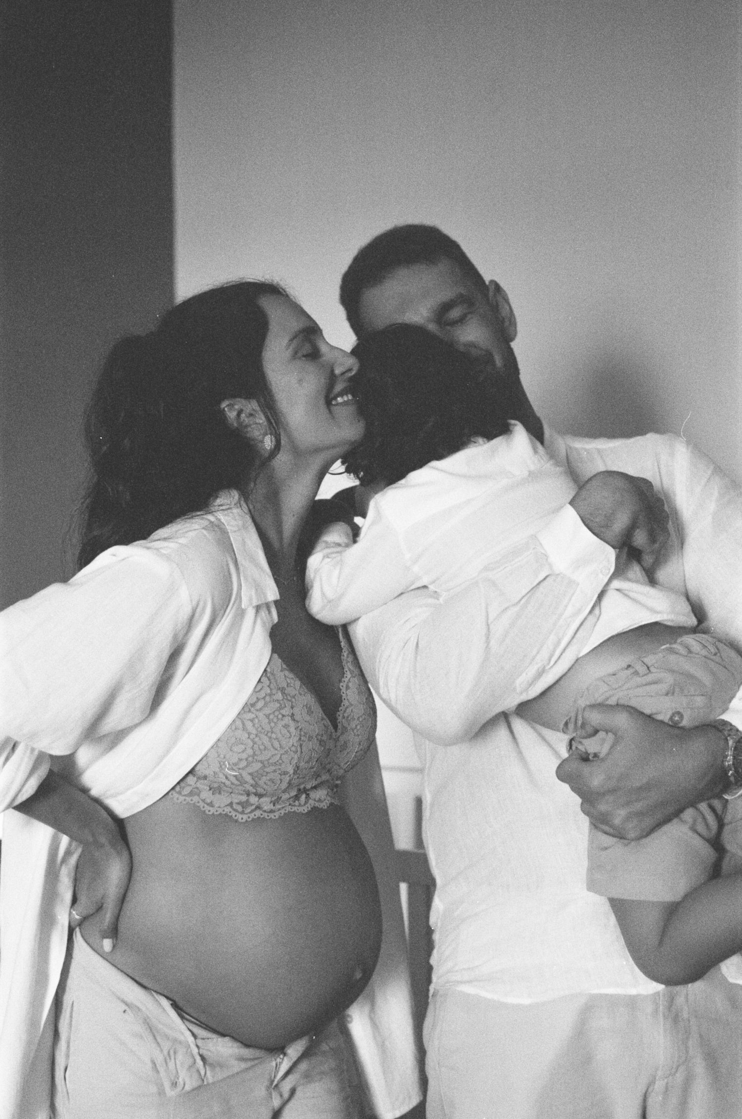 Black and white photo of a pregnant woman and man kissing while holding a newborn baby and embracing a child.