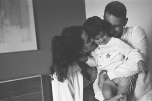 Black and white photo of a loving family, parents smiling and embracing their young toddler child indoors.