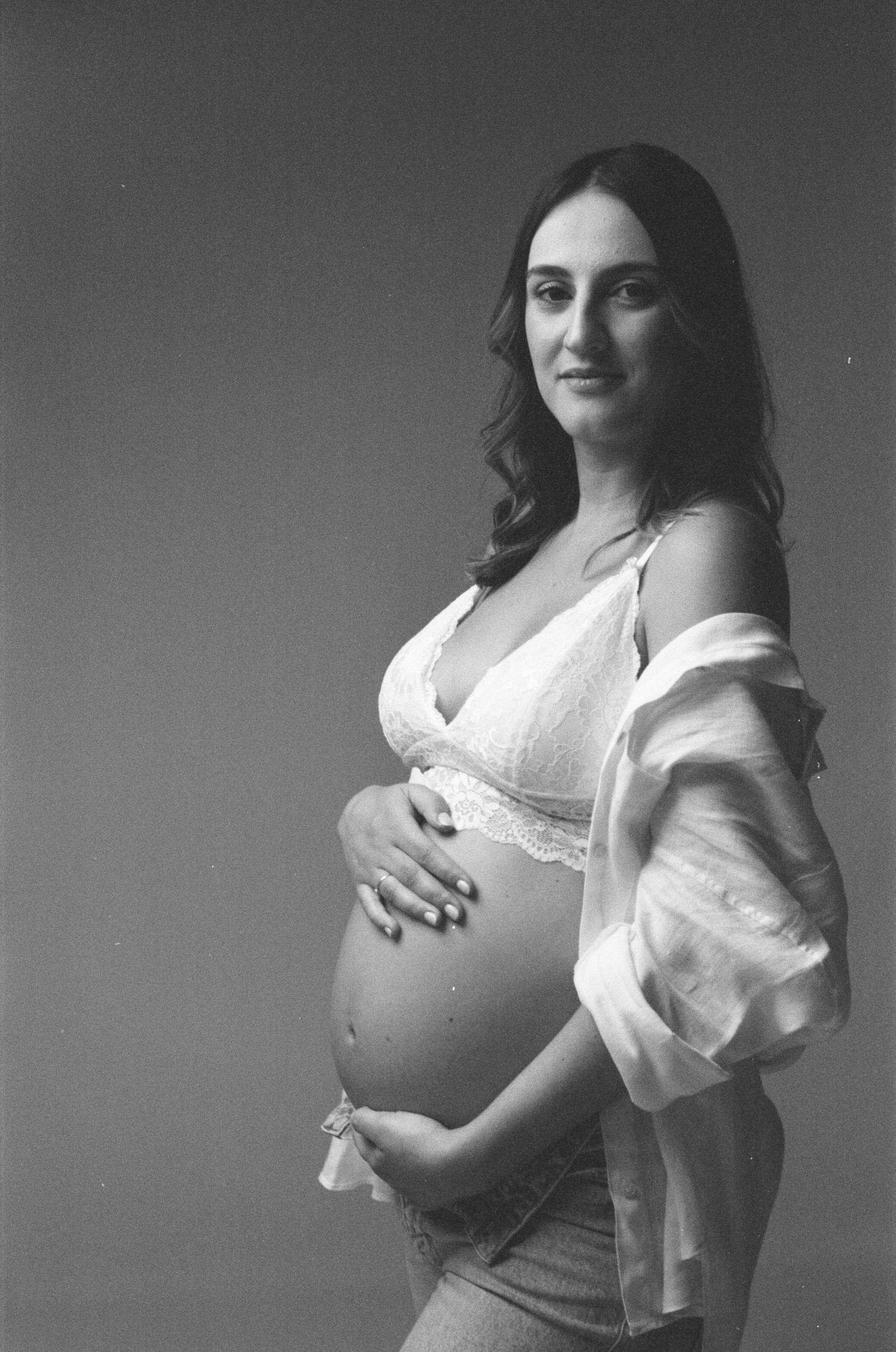 Black and white maternity photo of a pregnant woman in lace bralette cradling her baby bump in a studio.
