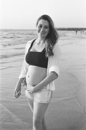 Smiling pregnant woman in black crop top and white shirt poses on sandy beach in black and white photo.