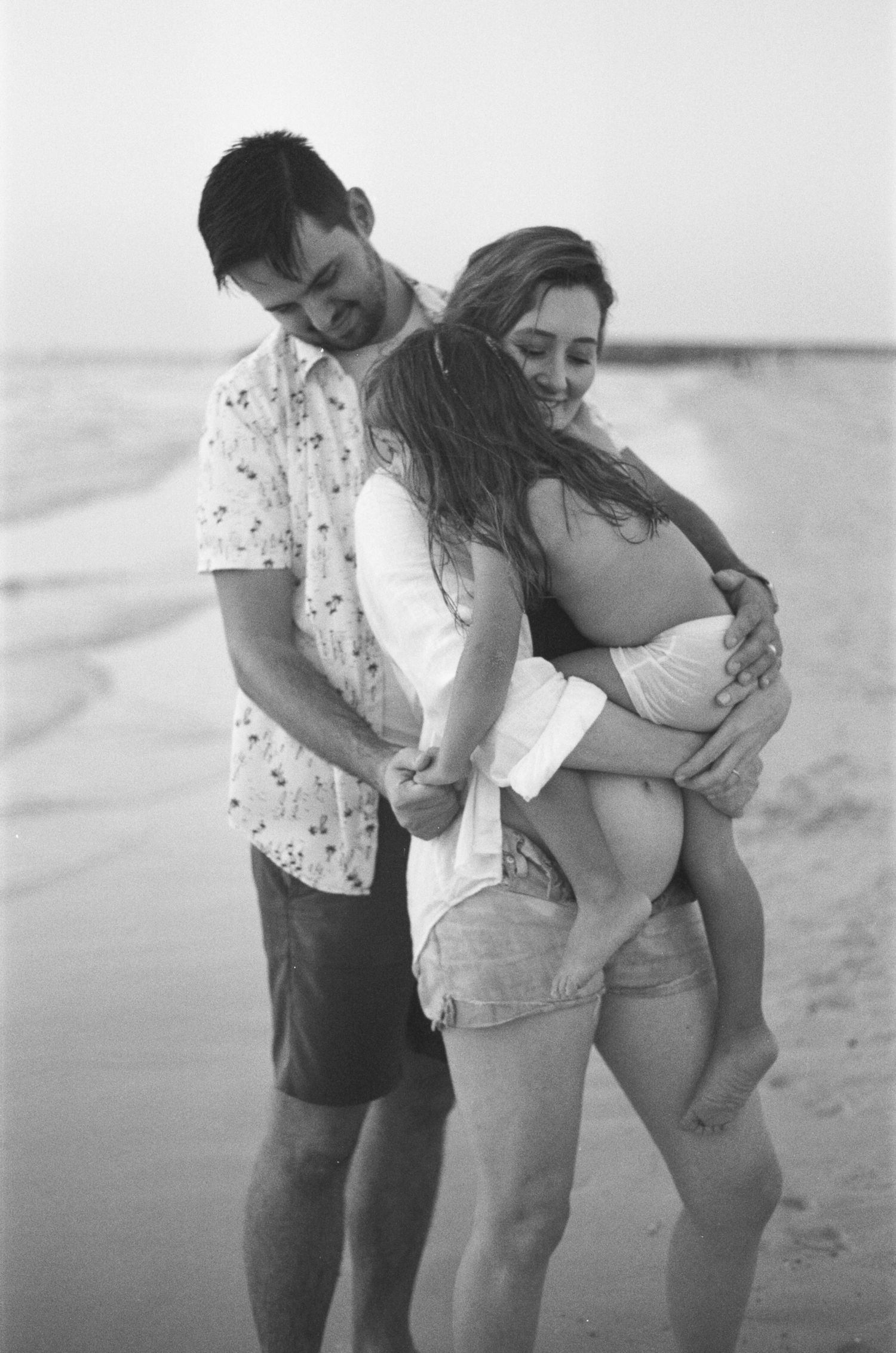 Black and white beach photo of a couple embracing, man holding pregnant woman's belly on sandy shore.