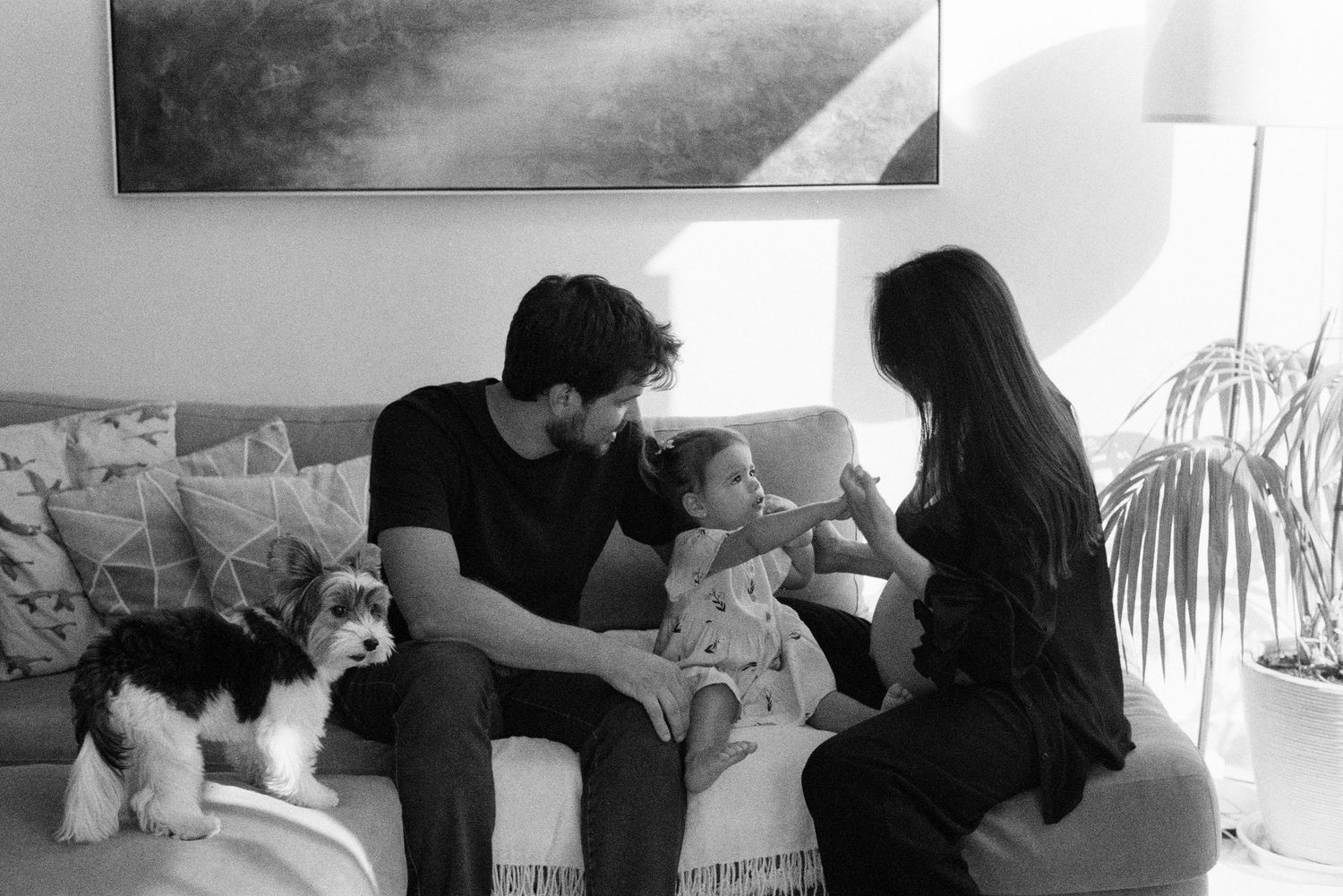 Black and white family photo of parents with toddler on couch, fluffy dog beside them in cozy living room.