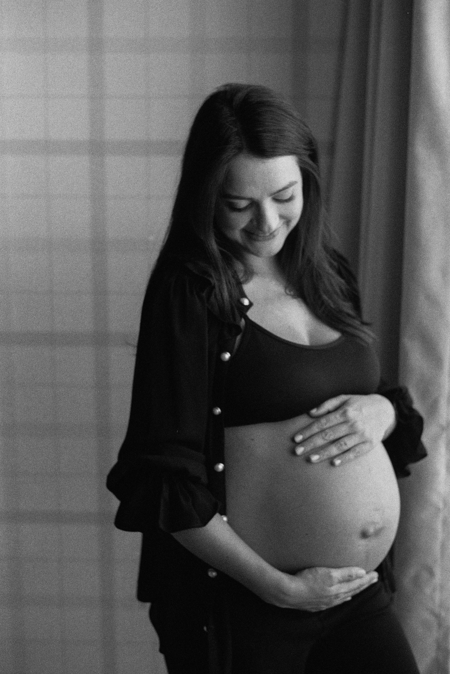 Black and white maternity photo of a smiling pregnant woman cradling her bare baby bump near a window.