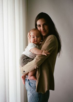 A young mother with dark hair tenderly holds her baby near a bright window, both dressed casually in neutral tones.