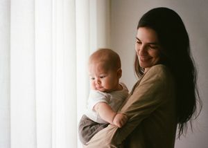 A smiling young mother with long dark hair holds her infant baby near a bright window with white curtains.