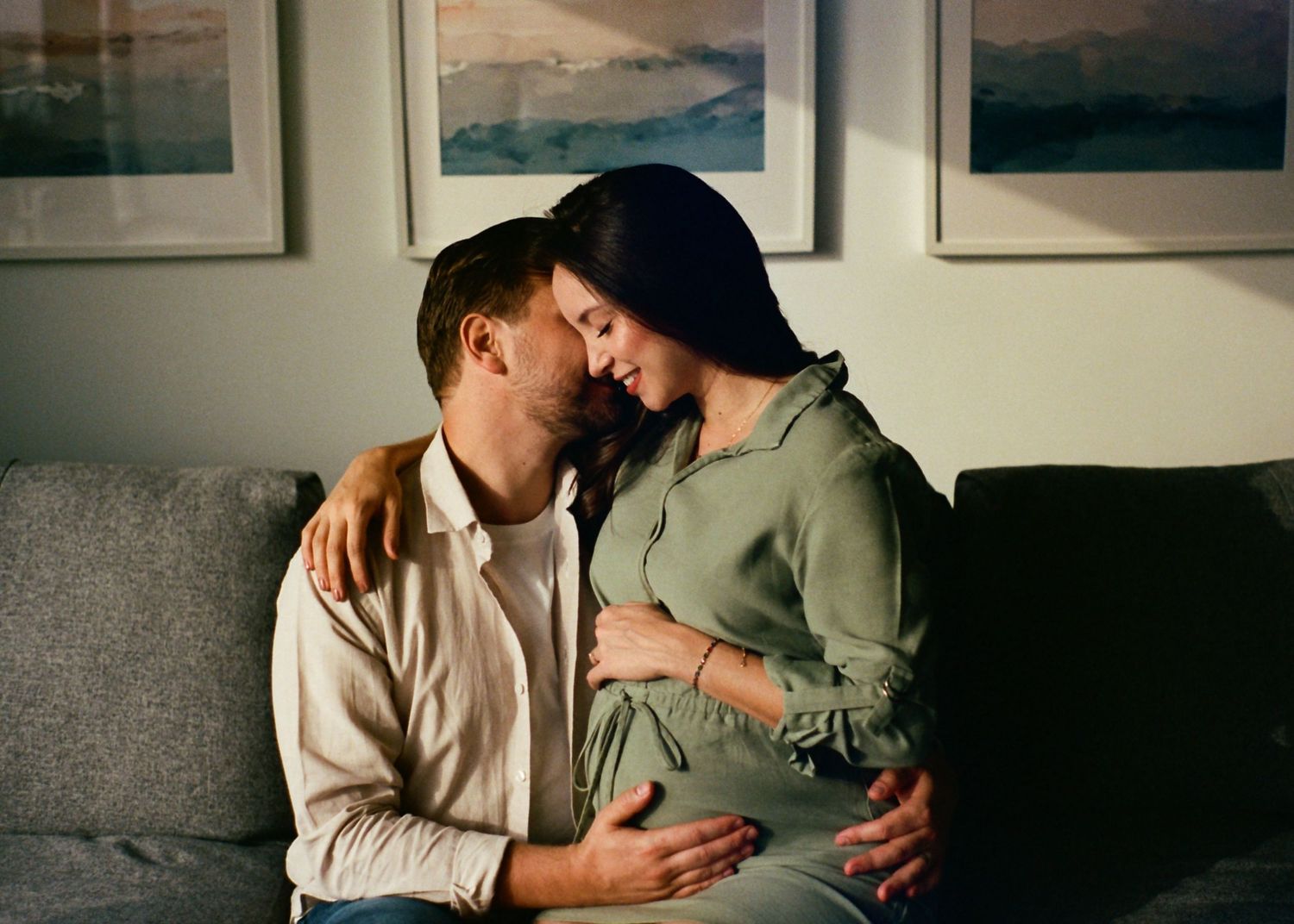 Couple sharing a tender moment on a couch, pregnant woman in green dress with partner embracing her baby bump.