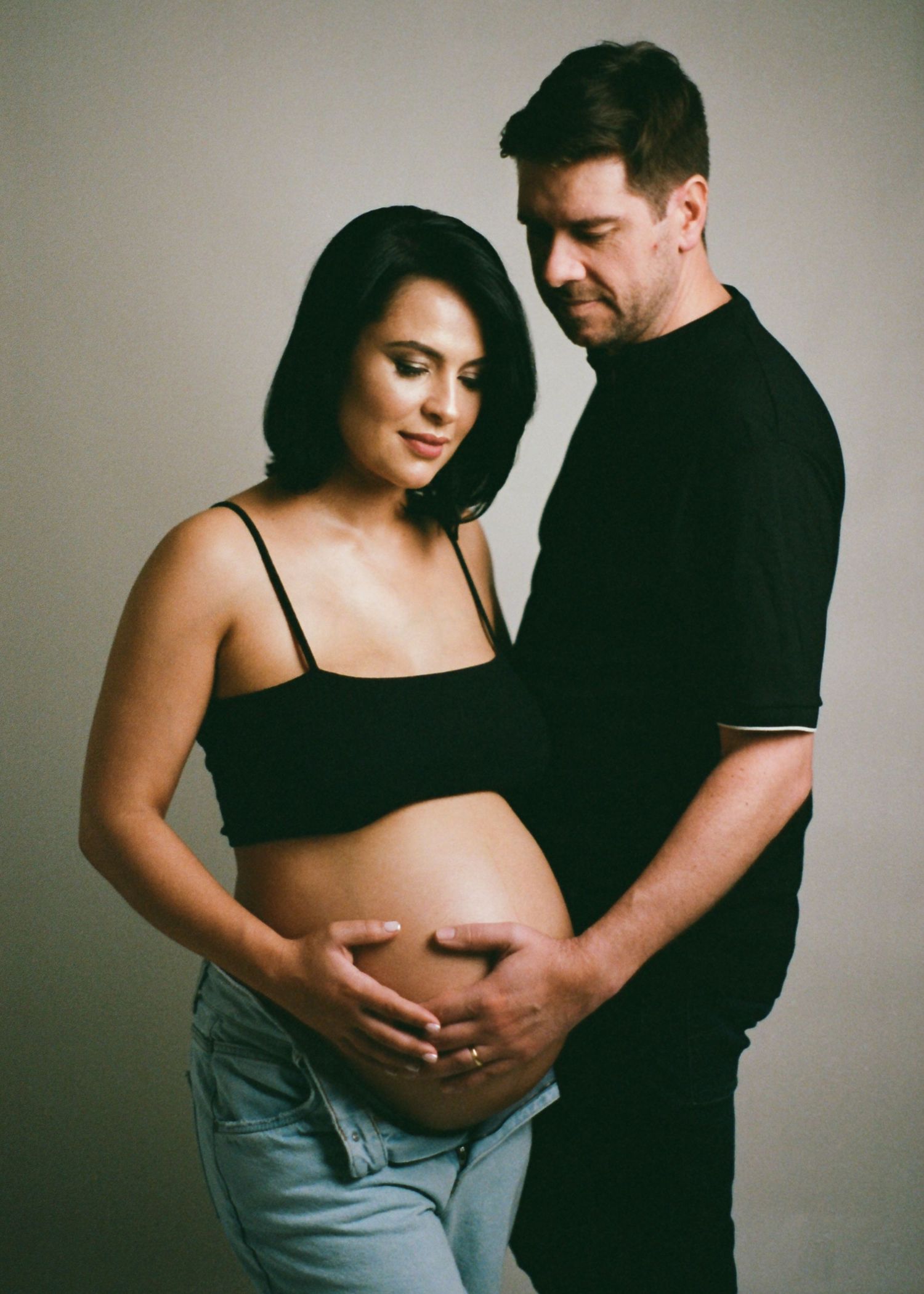 Couple sharing a tender maternity photo shoot moment, with man embracing pregnant womans baby bump against neutral background.