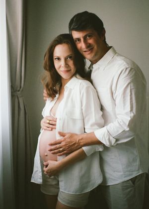 Expecting couple in white outfits posing together, man embracing pregnant woman showing baby bump, maternity photo session.
