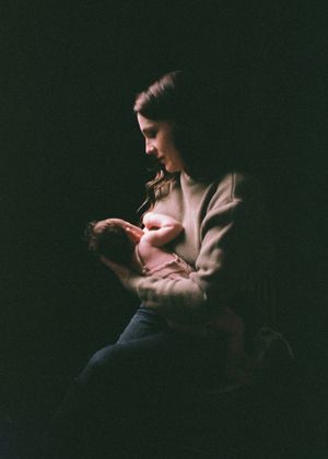 A mother tenderly holds her newborn baby in a dimly lit, intimate portrait captured on film photography.