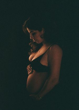 Pregnant woman in black bra cradling baby bump in dramatic dark studio maternity portrait photography.
