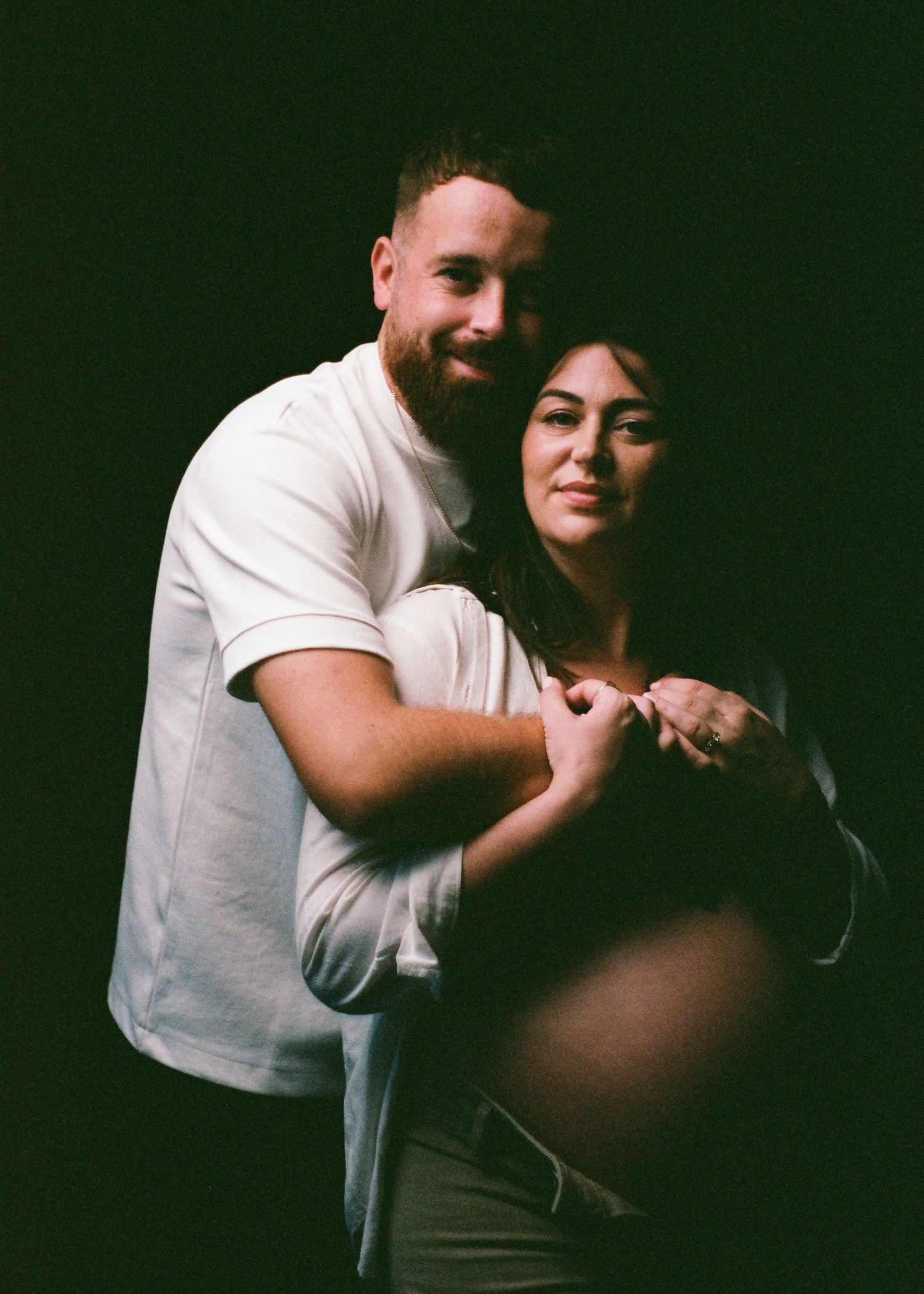 A couple in white shirts pose together for a maternity photo shoot against a dark background.