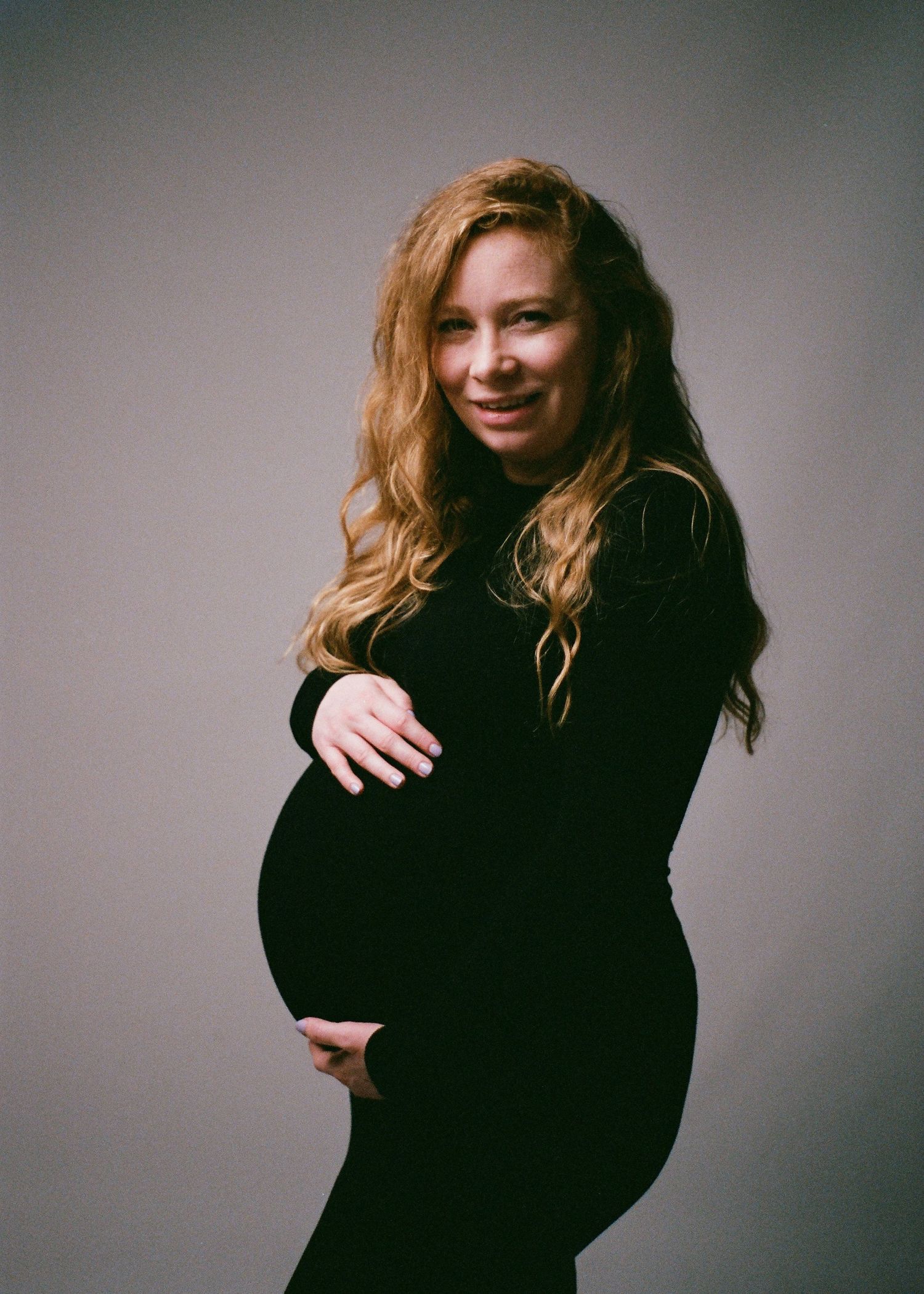 Pregnant woman in black dress cradling baby bump, smiling softly in a professional maternity studio portrait.