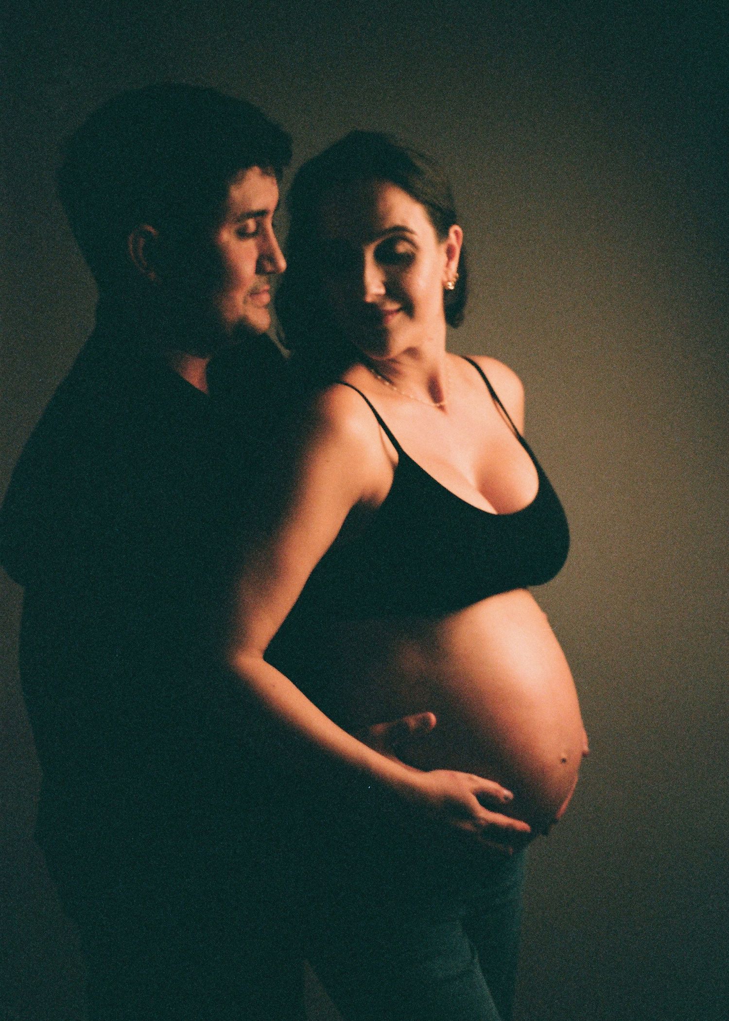 A couple poses for a maternity photo, the pregnant woman cradling her bare belly while her partner embraces her from behind.