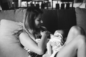 Black and white photo of a young woman sitting on a couch tenderly holding and gazing at a newborn baby.