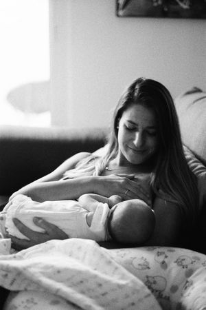 Black and white photo of a young mother breastfeeding her newborn baby while sitting on a couch at home.