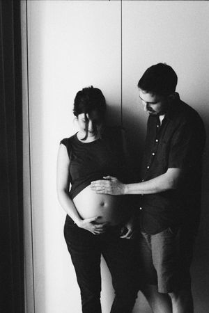 Black and white maternity photo of a couple tenderly touching a pregnant belly against a light wall.