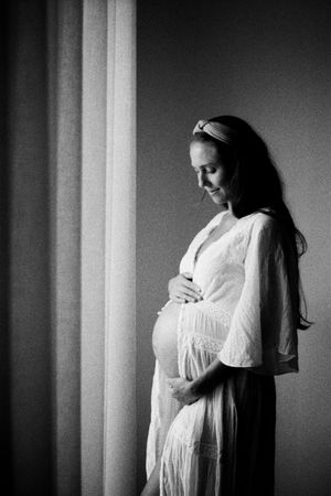 Black and white maternity photo of pregnant woman in white dress cradling baby bump by a window.