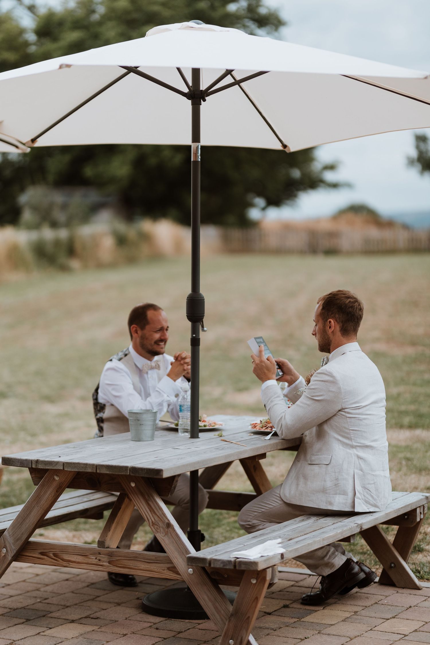 Two grooms in suits dining outdoors at a picnic table under a white umbrella on a sunny day.