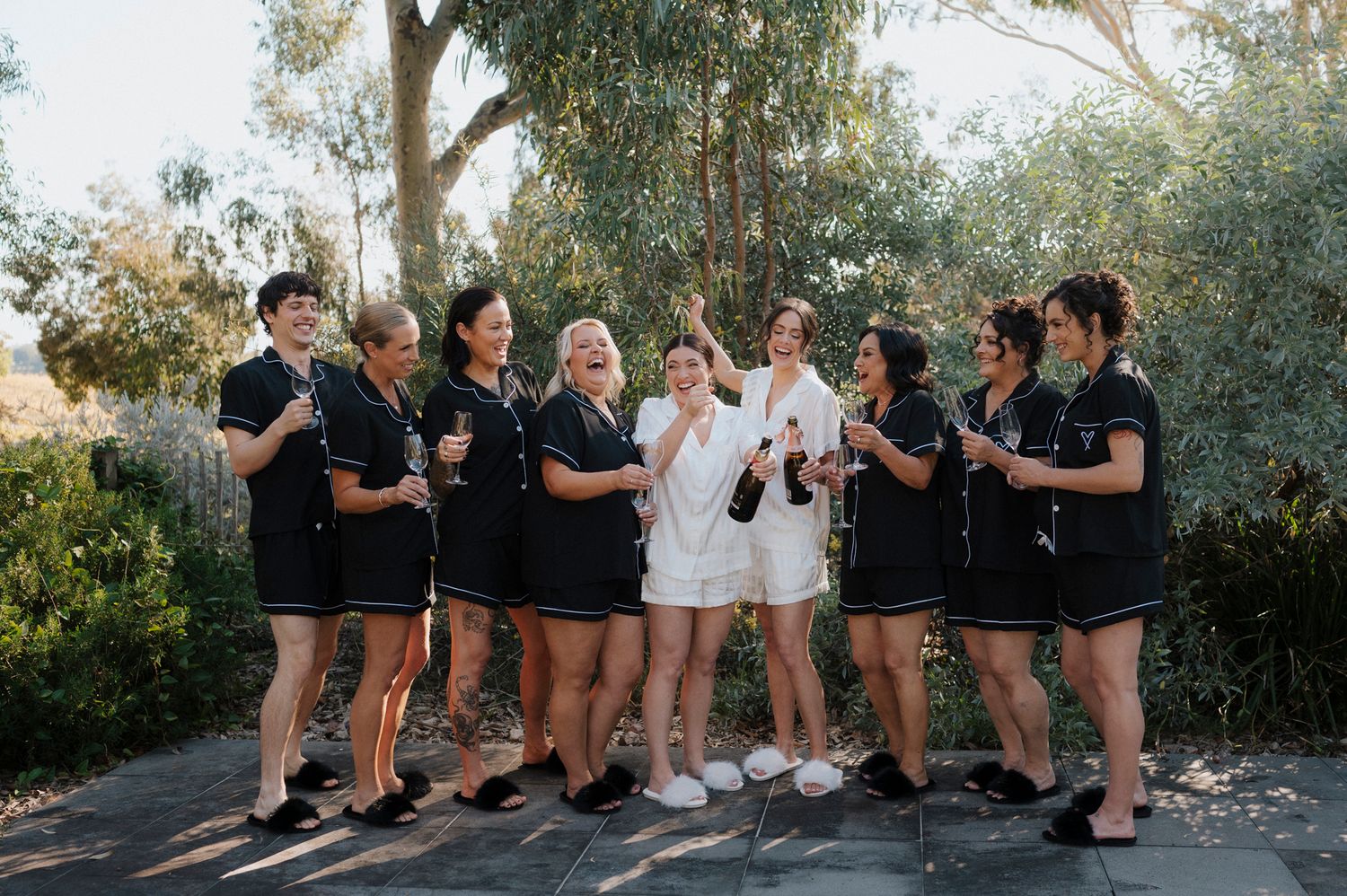 Bride in white romper celebrates with bridesmaids in matching black outfits, popping champagne outdoors.