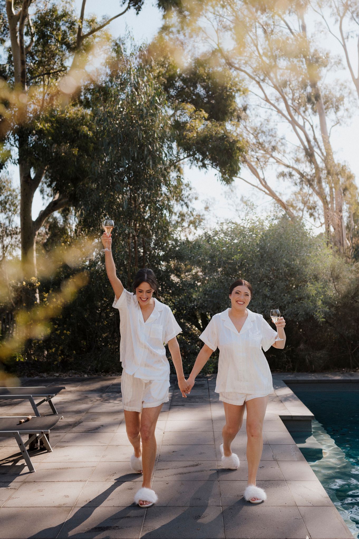Two women in matching white pajamas holding wine glasses by a pool surrounded by eucalyptus trees.