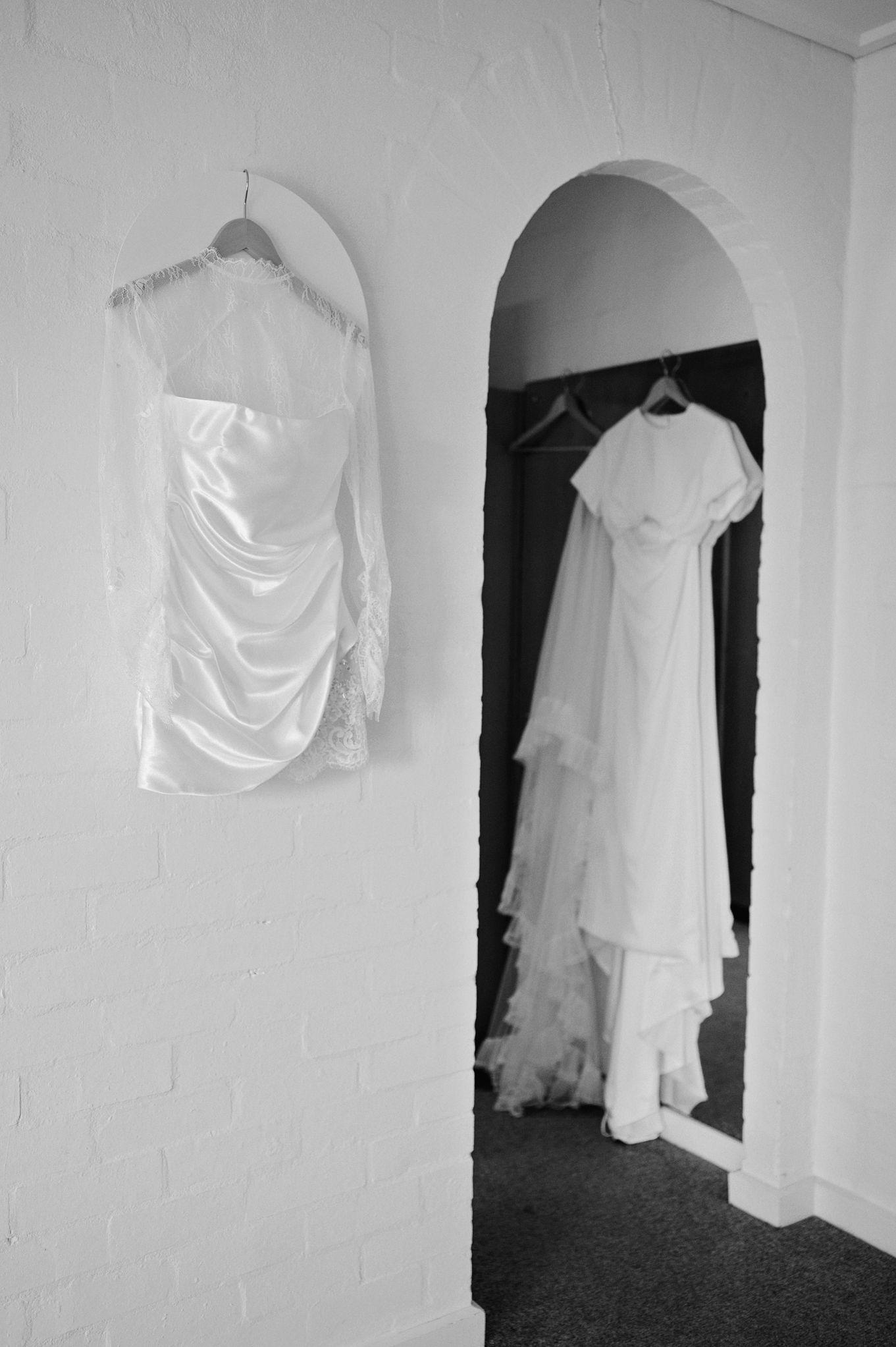 Black and white photo of two elegant white wedding dresses hanging in a bridal boutique with arched doorway.