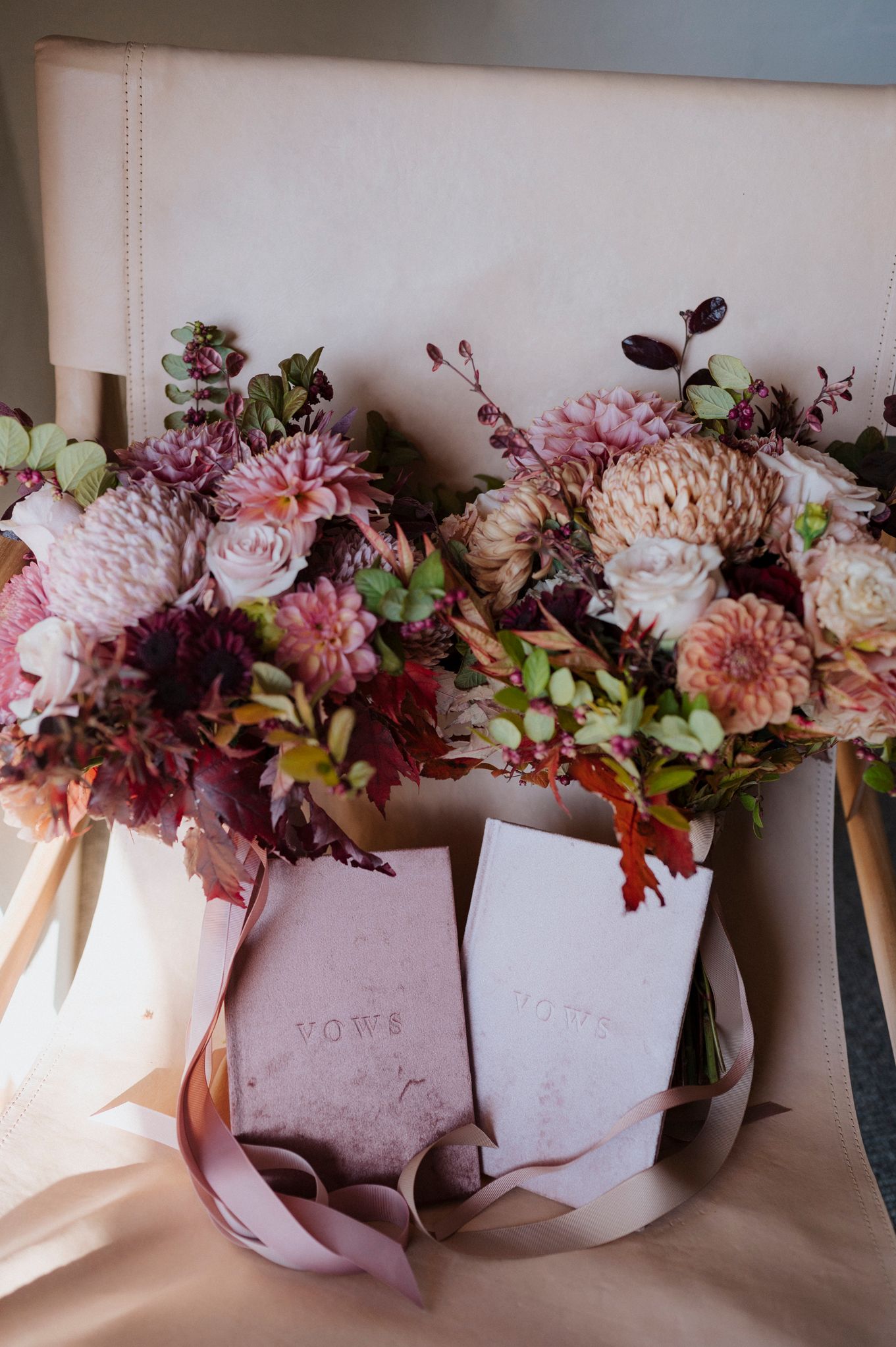 Two lush autumn bridal bouquets with dahlias and roses in mauve and burgundy tones, paired with elegant vow books on a chair.