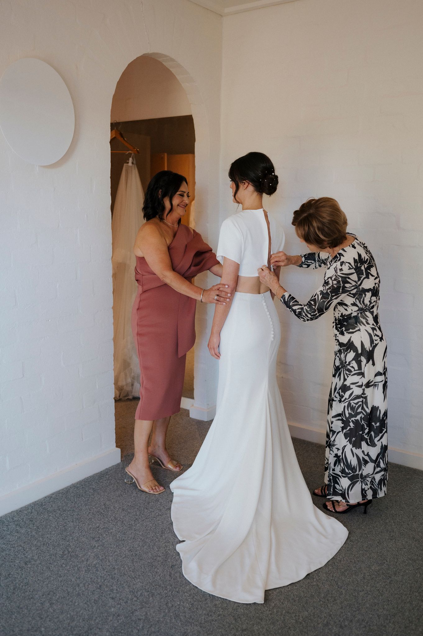 Bride in white gown being helped by two women in bright room before wedding ceremony.