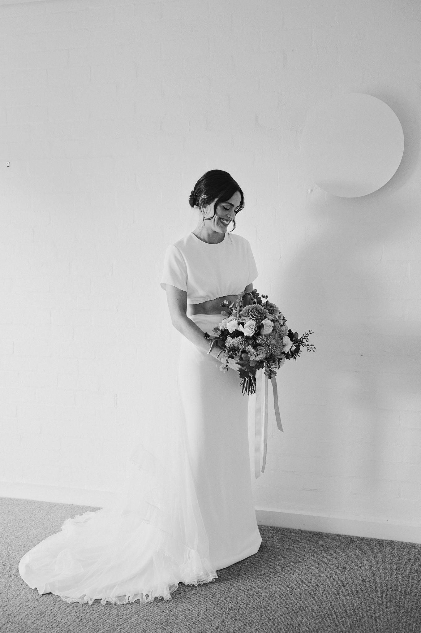 Bride in elegant white wedding gown with short sleeves holding a floral bouquet, smiling in a minimalist black and white photo.